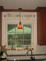 An Italian kitchen wall quote above the kitchen window and in between the kitchen wall cabinets, with kitchen sink and a pendant