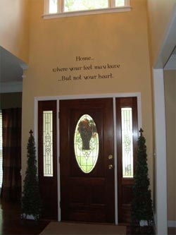 Home..where your feet may leave...But not your heart. A wall decal in between the window and the main entrance door with 2 indoor plants on both sides of the door.