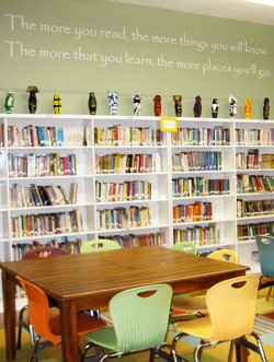 An inspirational wall decal in the library room with books in the shelves and a table with chairs - the more you read, the more things you will know. The more that you will learn, the more places you will go.